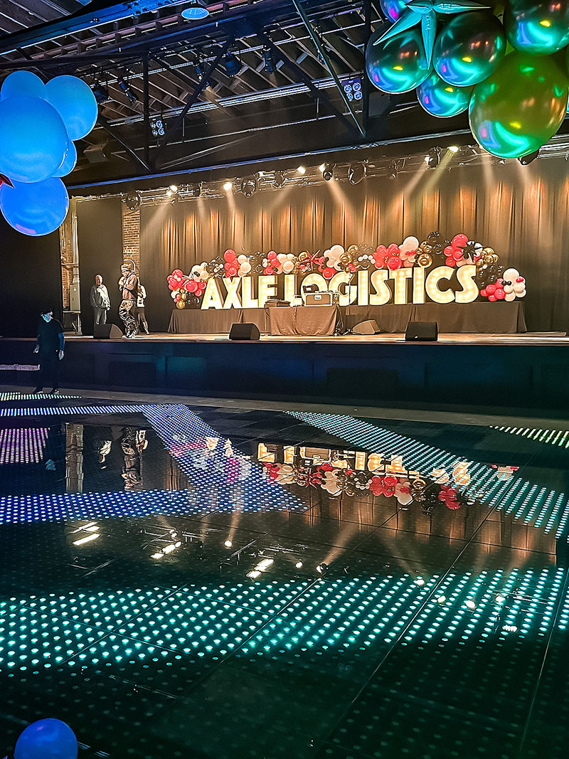 large illuminated letters spell Axle Logistics surrounded by metallic colored balloons and an LED dance floor