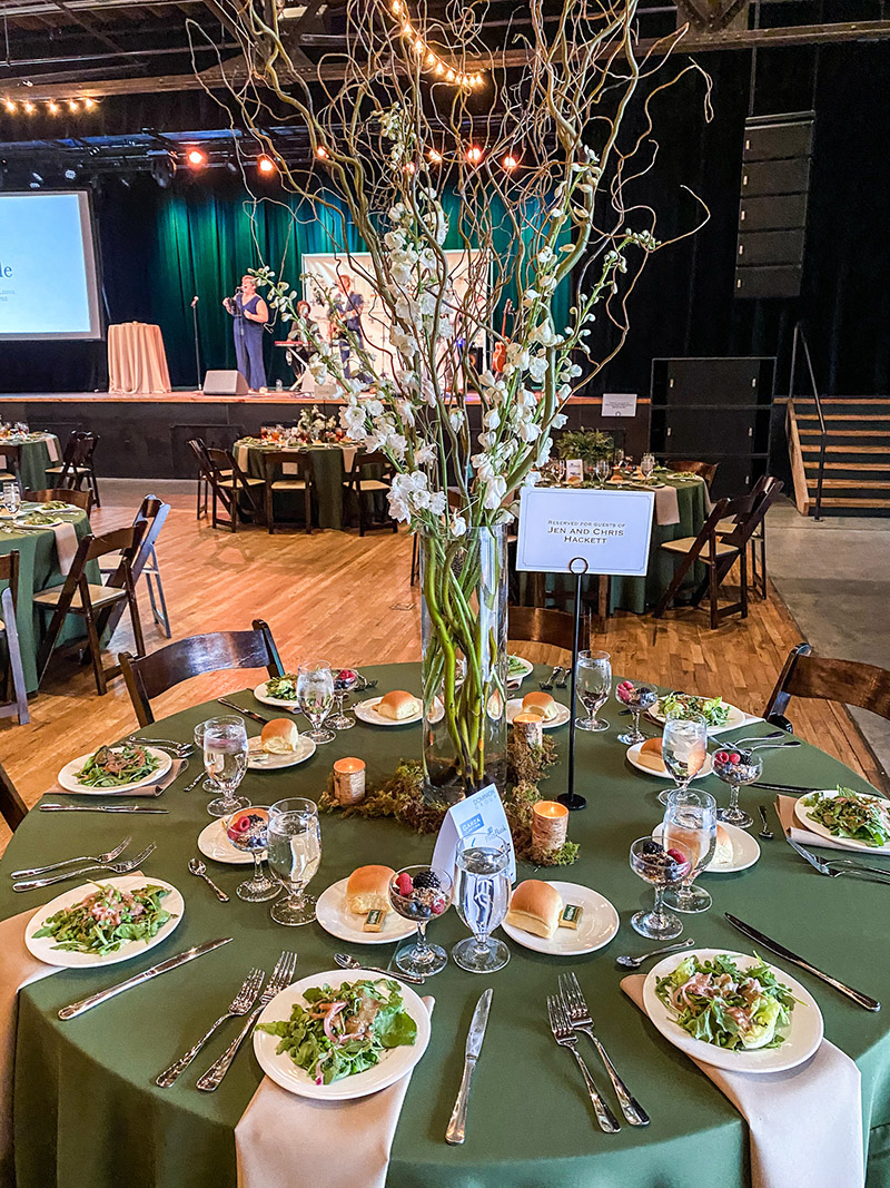 tables set with salad and dramatic floral centerpieces for a non-profit fundraiser with people standing on stage in the background