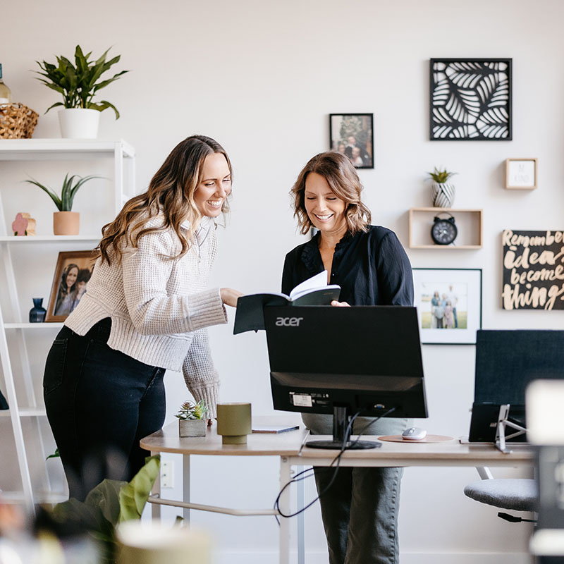 two women look at a book together while standing in a modern office with bright natural light